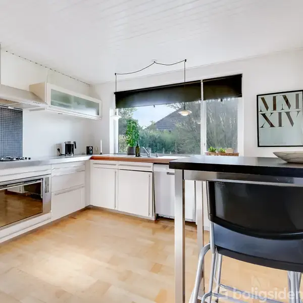 Et moderne køkken med hvide skabe og stål emhætte. Et spisebord med sorte stole står i forgrunden. Vinduet viser grønne omgivelser, og væggen har en plakat med "MMXIV".