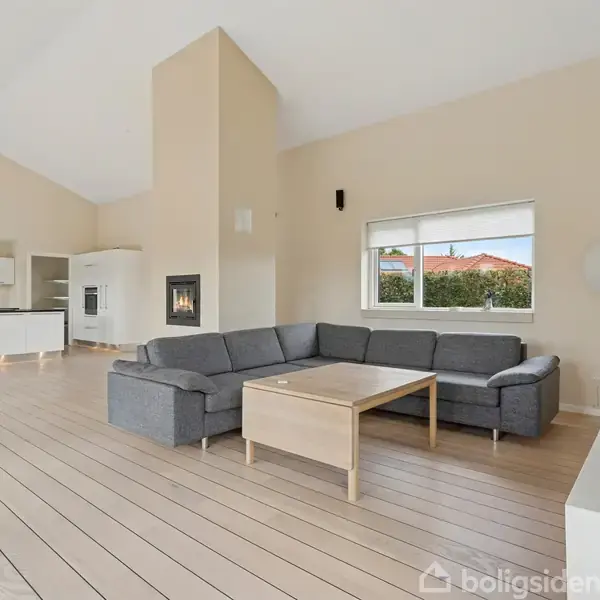 Stue med en grå hjørnesofa omkring et træbord. Rummet har lyse trægulve, store vinduer med udsigt til en hæk, og i baggrunden ses et moderne hvidt køkken.