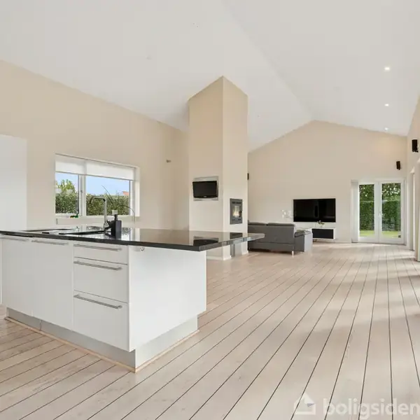 En moderne køkkenø står centralt i et åbent, lyst rum med lyse trægulve og store vinduer. En stue med sofa og tv er i baggrunden.