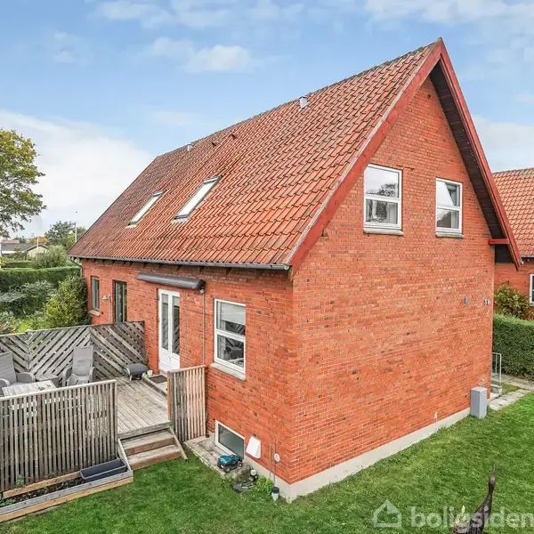 Rødt murstenshus med skråt tag og flere vinduer står midt i en have. En træterrasse med gelænder er ved siden af huset, omgivet af grønne buske og græsplæne.