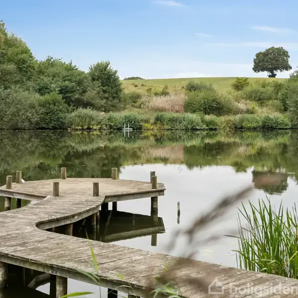 En træbro snor sig ud over en rolig sø med spejlinger, omgivet af frodig vegetation og bakker i baggrunden, under en klar himmel.