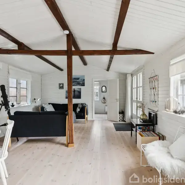 Et lyst stueområde med hvide vægge og træloftsplanker har en sort sofa, spisebord og stole. Et teleskop står ved vinduet, og dør fører indtil et andet rum.