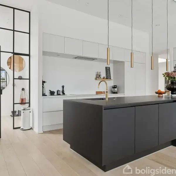 Moderne køkken med sort ø-køkkenbord, placerede stole og hængende lamper. I baggrunden ses hvide skabe, en dør med glasruder og minimal dekor. Gulvet er i lyst træ.