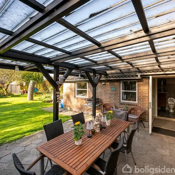 Terrassebord med stole, dækket af gennemsigtig tagkonstruktion, står på stenbelægning ved siden af murstenshus. Grøn have strækker sig ud i baggrunden med træer under klar himmel.