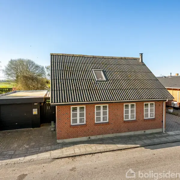 Rødt murstenshus med skråt sort-tag, flere vinduer og en tagkvist. Ved siden af ligger en garage. Husene har belægning af asfalt og fliser omkring. Omgivet af andre huse og grønne træer.