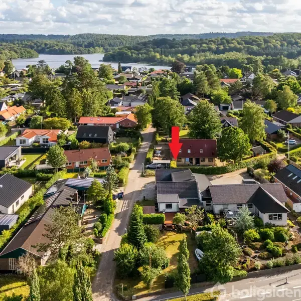 En rød pil peger på et hus i et boligkvarter omgivet af andre huse og frodig vegetation. I baggrunden ses en sø og skovklædte bakker under en blå himmel med skyer.