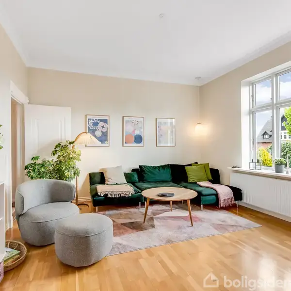 En stue med grøn sofa og lavt sofabord står på et gulvtæppe. Hylder med bøger og planter flankerer rummet. Store vinduer til venstre slipper naturligt lys ind.