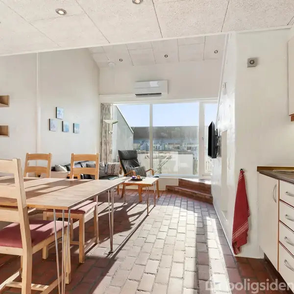 Spisestue med træbord og seks stole, sollys strømmer gennem store vinduer. En køkkenvask til højre, hylder med pynt til venstre, og en altandør i baggrunden.