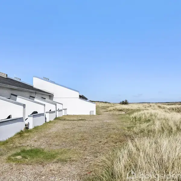 Række huse med vinklet tag står på en række. De grænser op til en eng dækket af tørre græsser under en klar blå himmel.