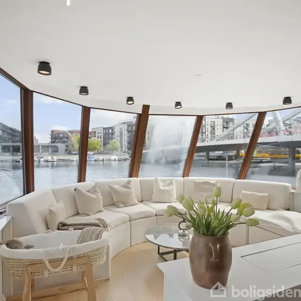 Stue med hvid hjørnesofa langs buede vinduer, udsigt over en bykanal og bro i baggrunden. Grønne planter og bordlampe skaber en moderne og hyggelig stemning.