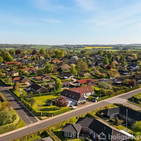 Beboelseshuse ligger spredt i en grøn, velordnet landsby omgivet af marker og træer under en skyfri blå himmel.