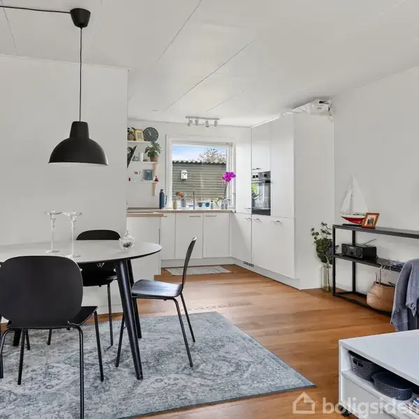 En moderne stue med sort spisebord og stole på et tæppe. Køkkenet ses i baggrunden. Et sofabord og en sofa står til højre, omgivet af dekorationer og lamper.
