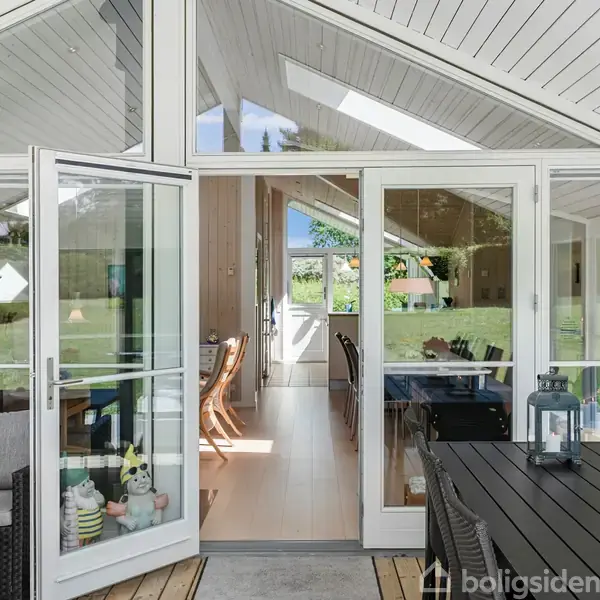 Glasdør åbner til lys stue med spisebord i elegant sommerhus. Indendørs pejs og udendørs terrasse med sorte møbler og røde blomster. Velholdt og moderne indretning omgivet af grøn natur.