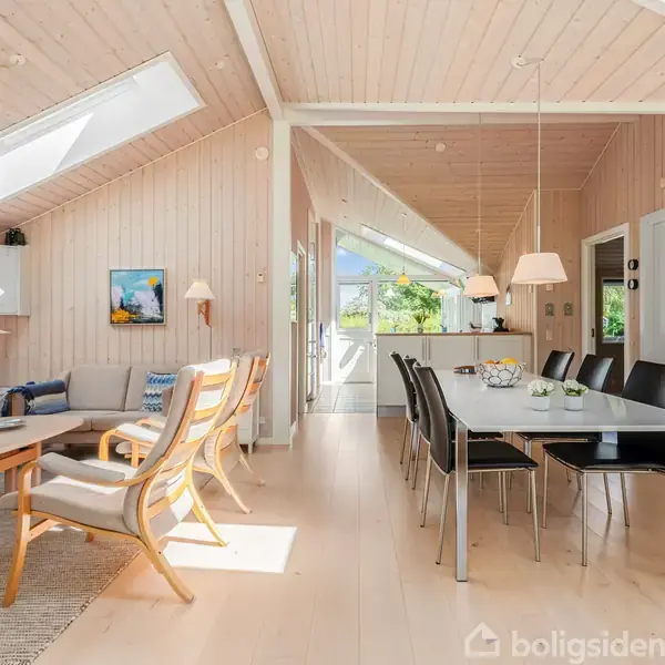 Stue med trævægge og loft, sollys skinner ind fra ovenlysvinduer, møbleret med sofagruppe til venstre og spisebord med stole til højre, dør fører til køkkenområde.