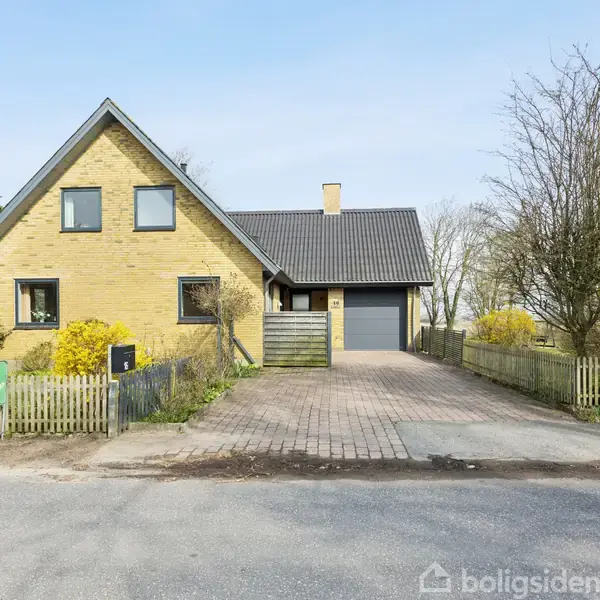 Gult murstenshus med trekantet gavl står foran indkørsel og hegn. Omkringliggende have har buske og træer. En blå "til salg"-skilt placeres til venstre for indkørslen.