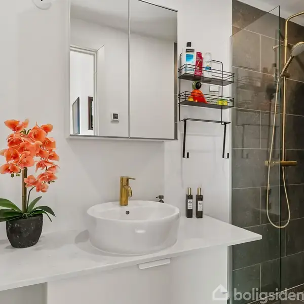 En moderne badeværelseskonsol med en rund vask og guldfarvet vandhane, flankeret af en plante med orange blomster. Baggrunden har et bruseområde med grå fliser og en vægmonteret opbevaringshylde.