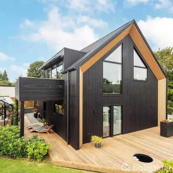 Sort, moderne hus med store vinduer står på en terrasse omgivet af grønne træer og planter. I baggrunden ses et drivhus og en have.