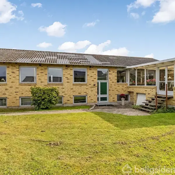 Et murstenshus med flere vinduer står stille omgivet af en grøn græsplæne, under en blå himmel med få skyer. Huset har en veranda med en overdækning.