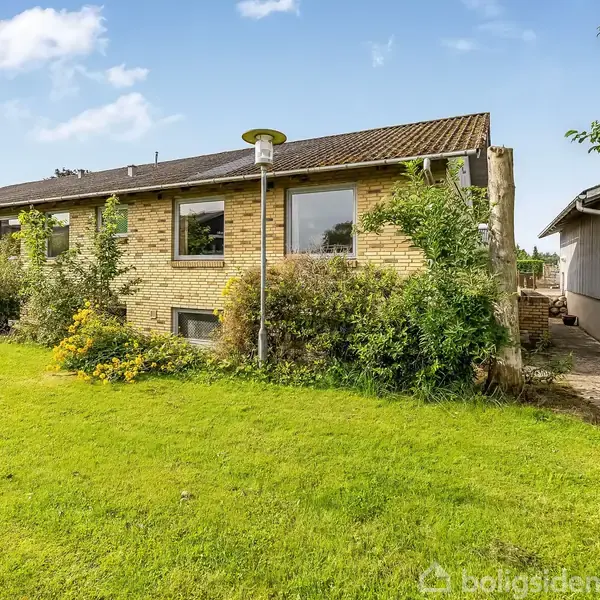 Et gult murstenshus med skråt tag og flere vinduer omkranset af frodige buske og græsplæne. Til højre ses en tilbygning eller garage ved siden af en flisebelagt gangsti.