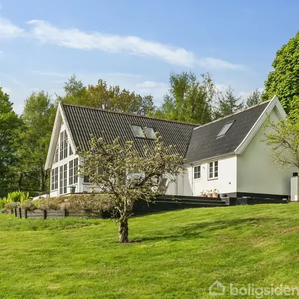 Hus med sort tag og store vinduer i en frodig have. Græsplænen er grøn, med træer omkring huset. En luftvarmepumpe er monteret ved siden af huset.