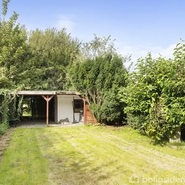 En carport står tom i en have, omgivet af frodige træer og buske. En græsplæne strækker sig fra forgrunden til bygningen i baggrunden.