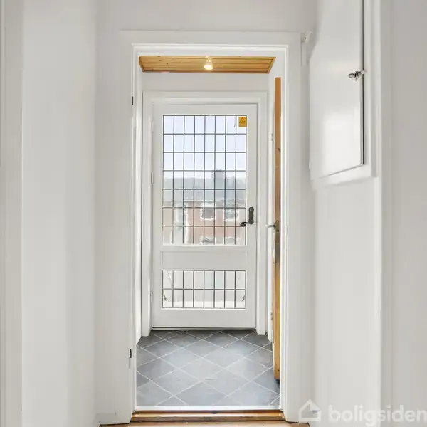 En hvid dør med glasruder leder ud til en balkon i en lys gang. Omkring døren er der hvidmalede vægge og en skifergrå flisebelagt gulv.