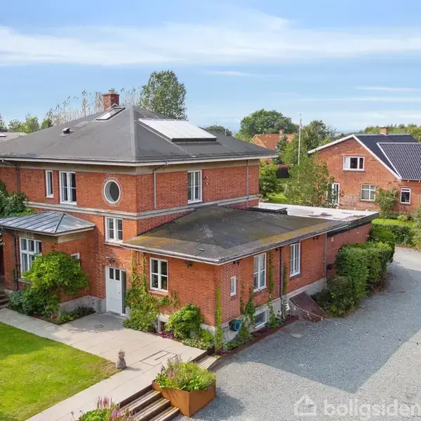 En rød murstensvilla med fladt tag står på en grusbelagt gårdsplads, omgivet af grønne buske og en velplejet have. Baggården har et moderne, fladt tagtilbygning.