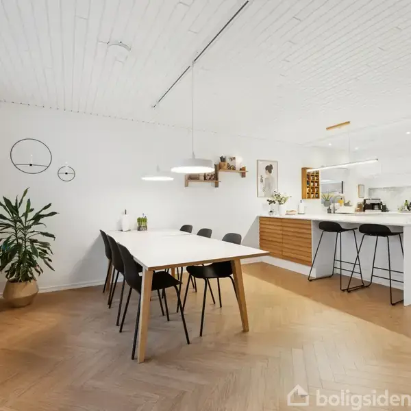 Lang spisebord med sorte stole arrangeret i en spisestue. I baggrunden ses køkken med hvidt interiør og barstole, lyse trægulve og loftslamper. Grøn plante ved vindueskarm.