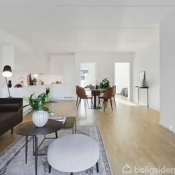 Stue med brunt sofaarrangement ved siden af et rundt sofabord, der har en plante i en vase. Stuen er i et lyst rum med spisebord og åben forbindelse til køkkenet.