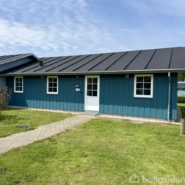 Et blåt træhus med sort tag står på en grøn græsplæne. En sti fører til hoveddøren i midten af huset, omgivet af buske og en klar himmel.