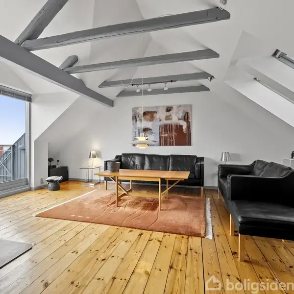 Rummelig stue møbleret med sorte sofaer rundt om et træbord på et tæppe. Skråvægge med ovenlys og fritliggende bjælker skaber loftshøjde. Store vinduer giver udsigt over en balkon.