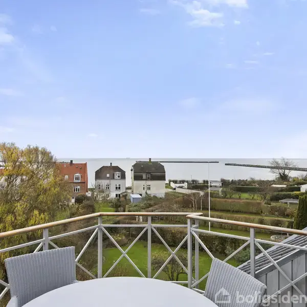 Et rundt bord med stole står på en altan. I baggrunden ses huse, en have og en udsigt over vandet under en skyfri himmel.