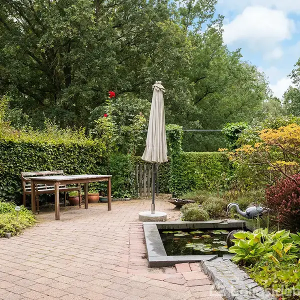 En have med en parasol ved et bord på en flisebelagt terrasse. Ved siden af er en lille dam omgivet af planter og blomster, omkranset af grønne træer og buske.