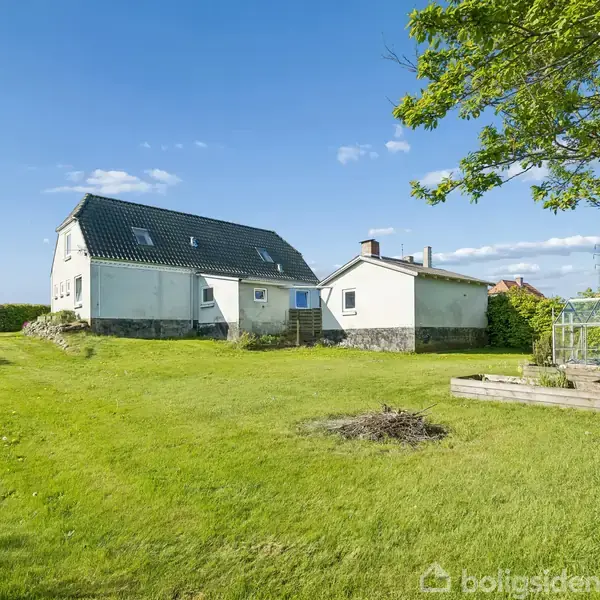 Et hvidt hus med sort tag står på en stor, grøn græsplæne. Til højre er der en terrasse og et drivhus. Himlen er blå med spredte skyer.