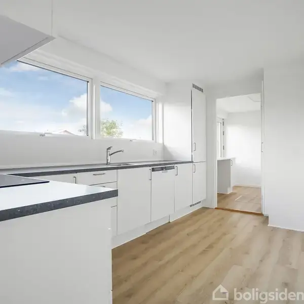Murstenshus med blå træbeklædning, beliggende ved en terrasse med fliser og grussti, førende til en grøn græsplæne. Omgivet af træer og buske under en blå himmel med skyer.