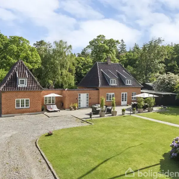 Rødt murstenshus med mørkt tegltag står stille i en grøn have med grusvej. Omkring er der tæt skov og blomstrende buske.