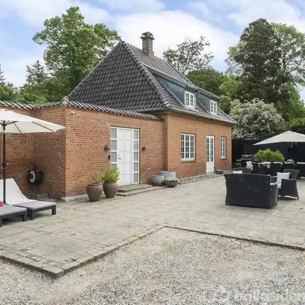 Rødt murstenshus med gråt tag står på en terrasse med solsenge og parasoller. Omkringliggende have med grønne træer og buske. Ingen tekst er synlig.