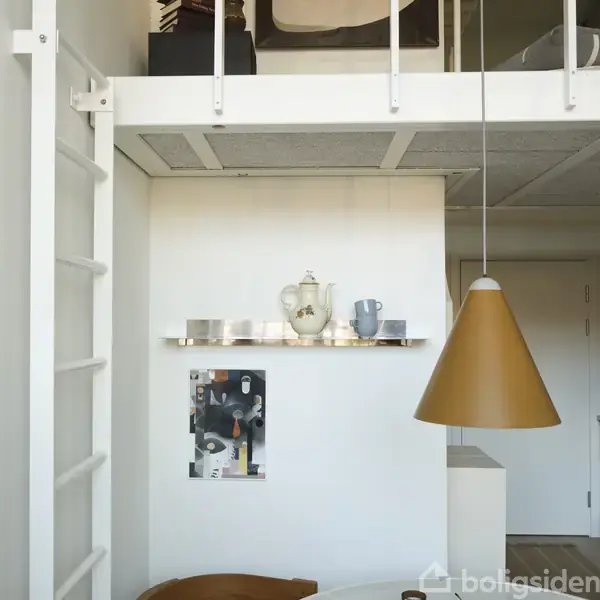 En hvid væg har en hylde med en tekande og kop. En plakat hænger nedenunder. En stor, brun lampeskærm hænger ned fra loftet.