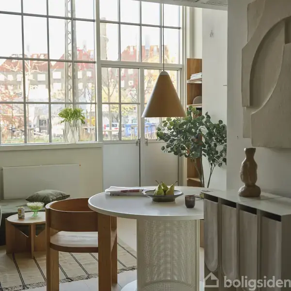 Rundt bord med frugtskål står i stue med store vinduer. Grøn plante og lamper skaber hyggelig atmosfære. Hylder i baggrunden med bøger. Udendørs bymiljø ses udenfor.