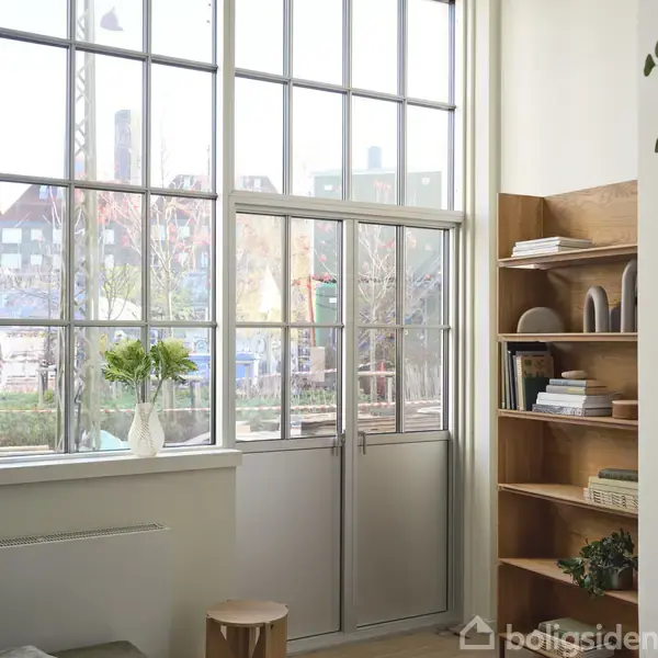 En glasdør med store ruder giver udsigt til en udendørs have. En vase med grønne blomster står på vindueskarmen, og en træreol med bøger er til højre.
