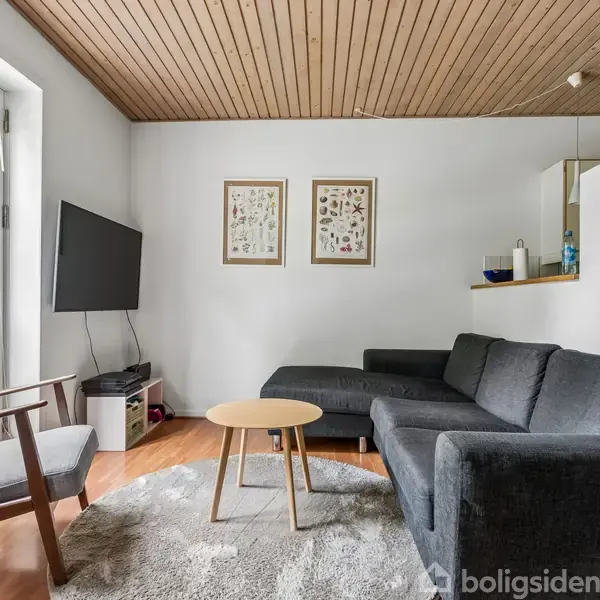 En grå sofa står i en moderne stue ved et rundt træbord. Et TV hænger på væggen overfor. Store vinduer bringer lys ind. To billeder hænger på en hvid væg.