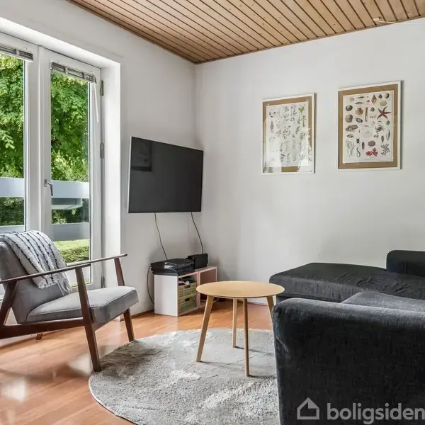 Stue med grå sofa ved siden af et rundt, træbord og en lænestol på et tæppe. Væggen har to botaniske plakater. Et TV hænger ved en fransk altandør med udsigt til træer.