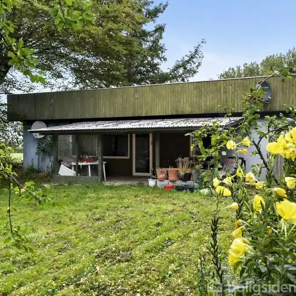 En etplans bungalow med fladt tag, omgivet af en frodig have med blomster og høje træer. Foran huset står der krukker og havemøbler på terrassen.