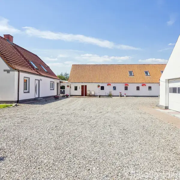 En gårdhave med flere hvidkalkede bygninger med røde tegltage omkring en grusbelagt gårdsplads, en garage med et kunstværk på væggen til højre, under en blå himmel.