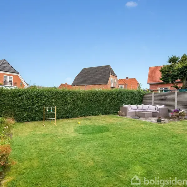 En velplejet græsplæne med et træ gyngestativ står i midten. Til højre er en terrasse med hjørnesofaer, omgivet af høje hække og huse med røde tage i baggrunden.
