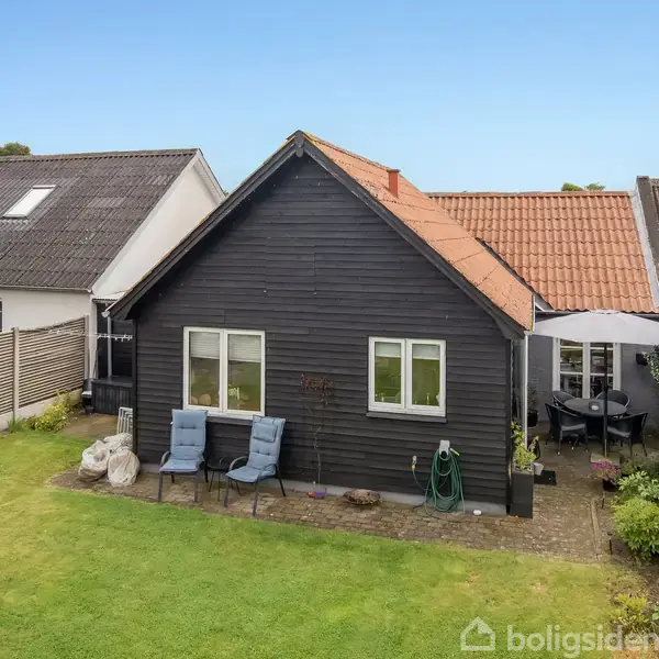 En sortmalet træhus står stille i en lille baghave med grøn græsplæne og terrasse. To blå stole er placeret nær husets væg ved siden af et rundt havebord.