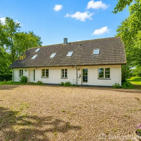 Hvidt hus med mørkt tegltag står på en grusplads omgivet af grønne træer under en blå himmel med enkelte skyer.