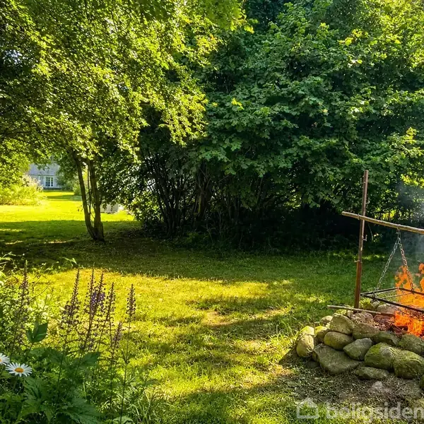Bål på jorden omgivet af sten, placeret i en frodig have med blomster og træer. I baggrunden ses en lysning med et hus.