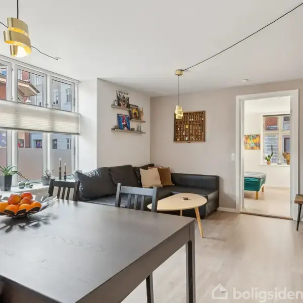 Stue med sort sofa mod væg, sofabord, og farverige puder; spisebord med frugtskål og stole; lys hænger fra loftet; lyse trægulve; vindue med persienne.