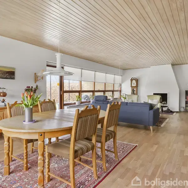 Spisebord med stole står i forgrunden, blomstervase på bordet. Stue med sofaer i baggrunden, stor vinduesvæg, brændeovn og dekorative elementer i hyggelig, lyst træbeklædt bolig.